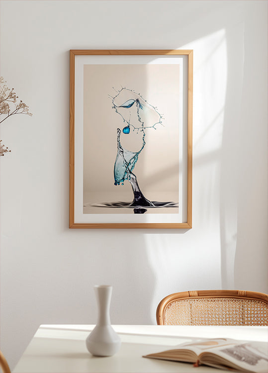 un'opera d'arte incorniciata di una goccia d'acqua sospesa a mezz'aria, con una combinazione di colori blu e bianco. L'opera d'arte è esposta su un muro bianco sopra un tavolo con un vaso bianco e un libro.
