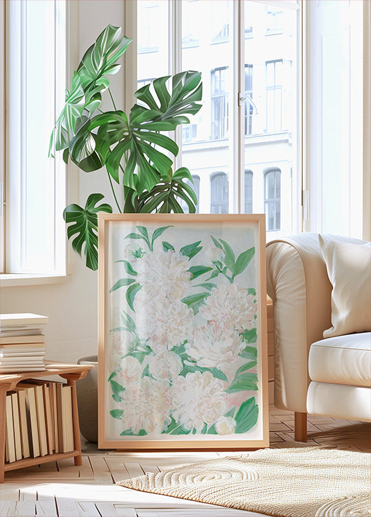una grande stampa incorniciata di fiori bianchi con foglie verdi, posta su un pavimento di legno in una stanza luminosa e ariosa con grandi finestre.